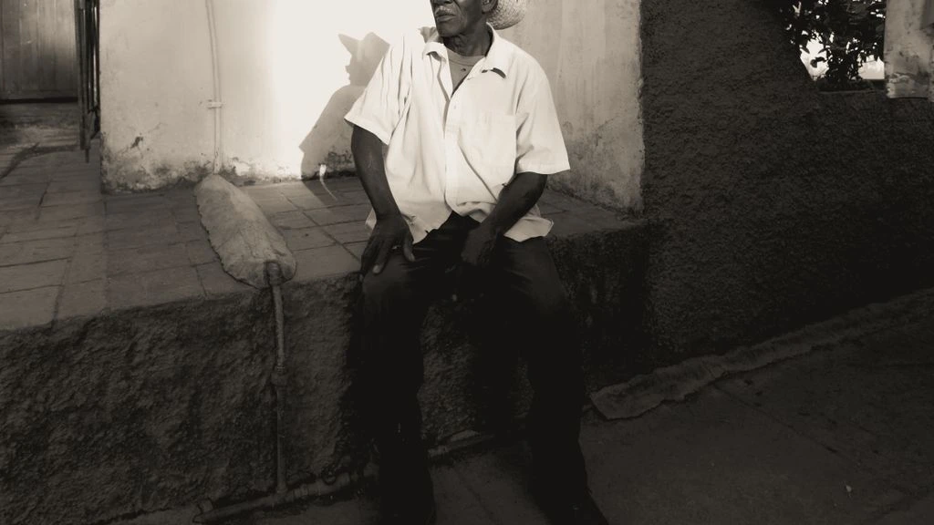 An older man sits outside a rebuilt stone house in a coastal village, late afternoon light, olive trees behind.